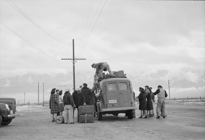 Man with pipe stands on top of bus loading luggage into rack, while others gather around.