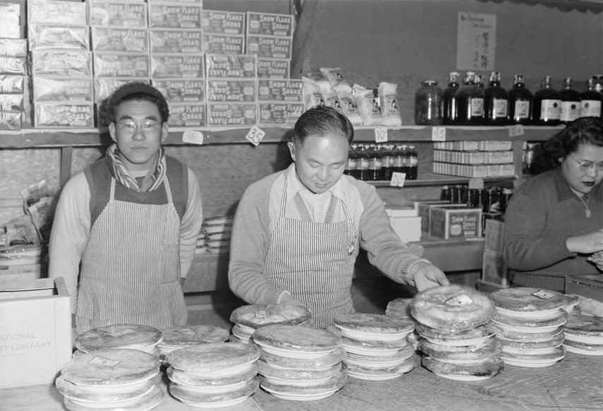 Two men and a woman standing behind store counter with display of pies.