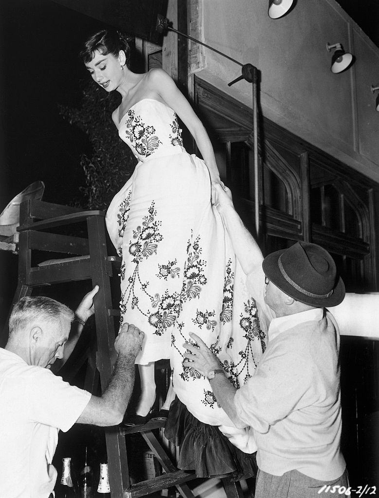 #40 Audrey Hepburn standing halfway up a lifeguard’s chair as she wears a flower-patterned dress.