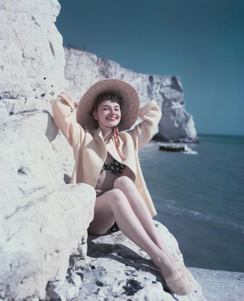 #56 Audrey Hepburn relaxes by the coast as she sits on a rock wearing a hat and beachwear, 1951.