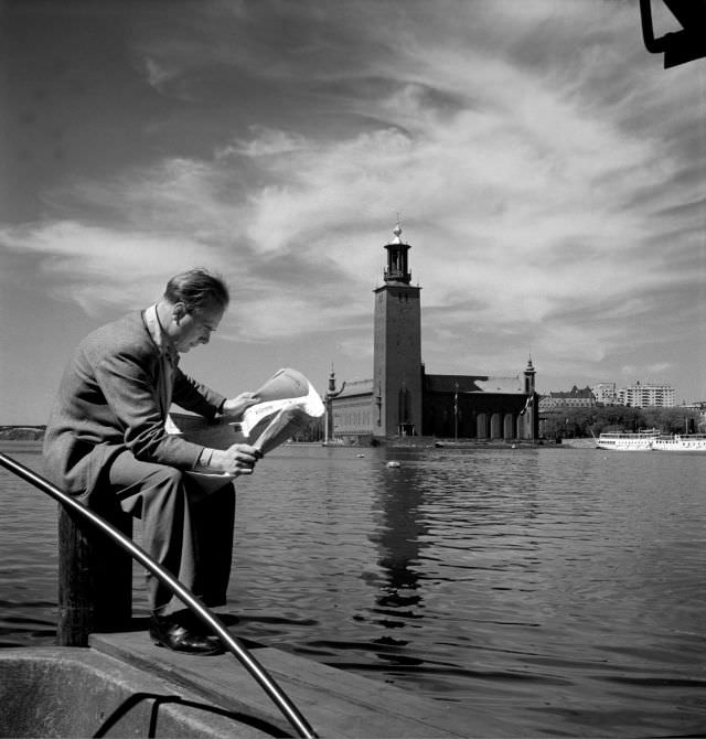 #6 A man sitting on a bollard and reading a newspaper. Stockholm City Hall and Riddarfjärden in the background.
