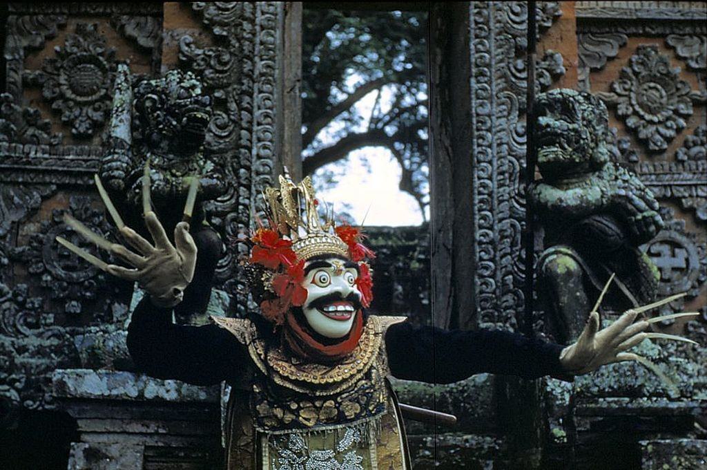 #23 A Balinese ‘topeng’ or masked dancer in ceremonial headdress stands in front of ornately carved doors, 1956.