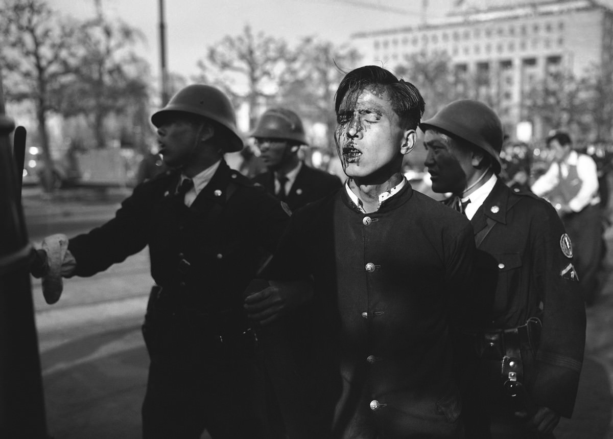 #13 A dazed Japanese youth, his face bruised and bleeding, is led from the riot scene by a policemen after pro-communist demonstrators were dispersed near the imperial palace grounds in Tokyo on May 1, 1952.