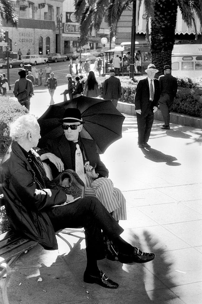 #34 Two men sitting on park bench sheltering from the sun under an umbrella, San Francisco California, 1970.