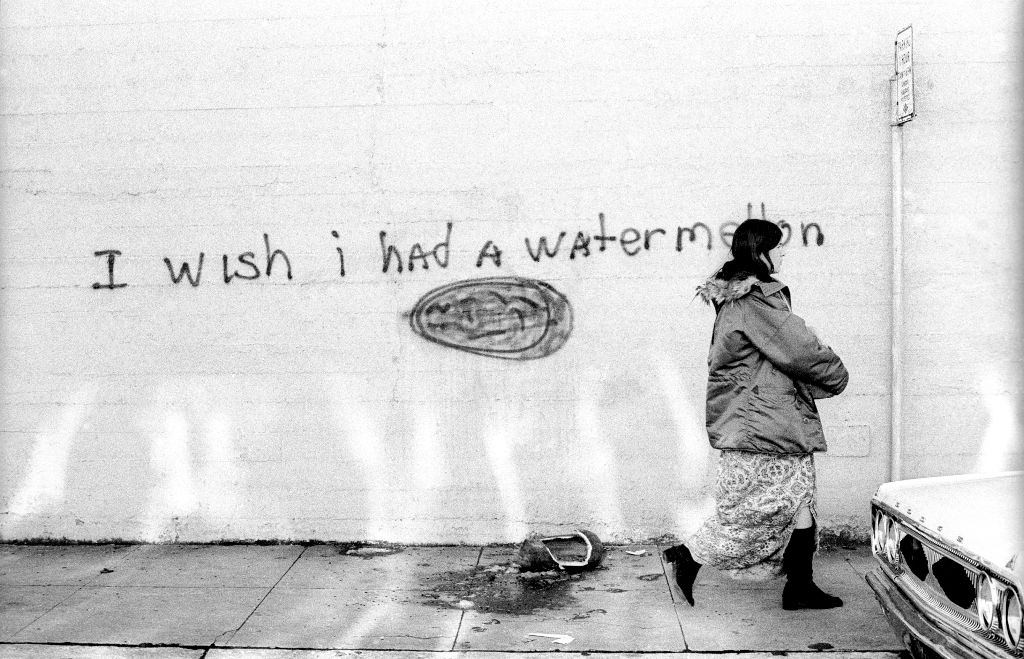 #74 A woman walking by a message that says “I wish I had a Watermelon” with a crushed watermelon lays on the ground circa January, 1972.