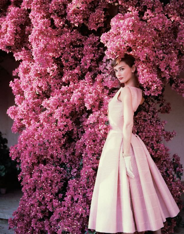 #17 Audrey Hepburn in a pink dress, standing next to a flowering bougainvillea plant, 1955.