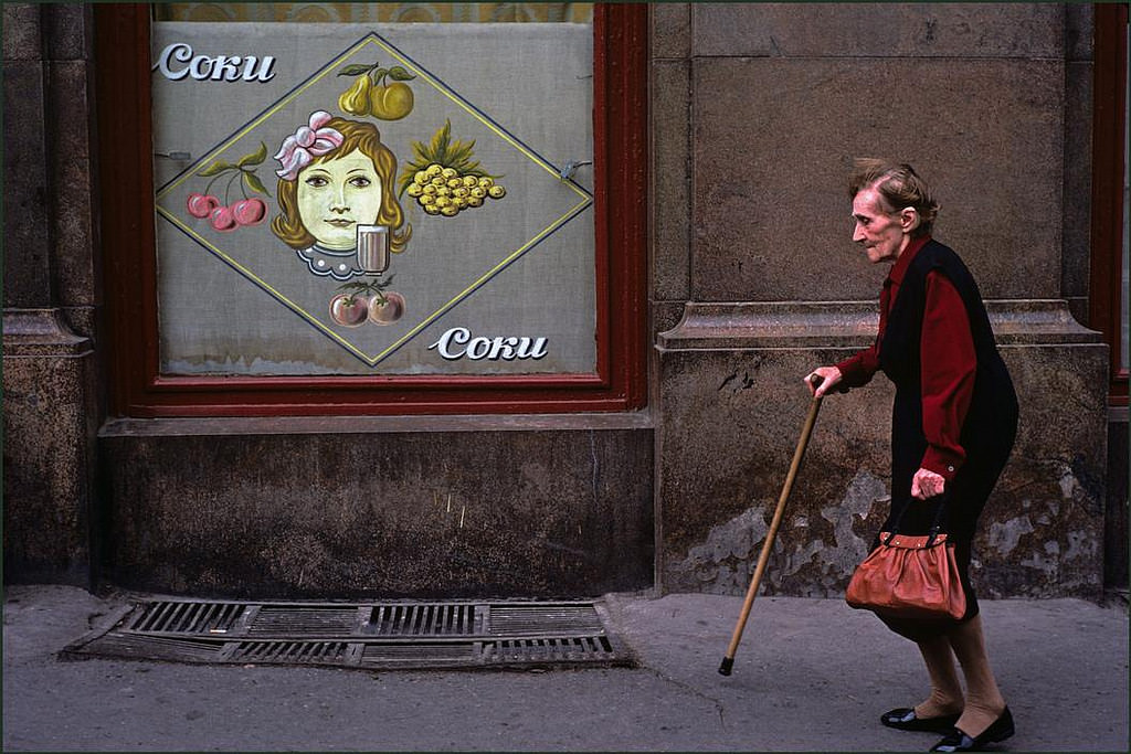 #24 Elderly lady with walking stick passing a window advertising a fruit drink.