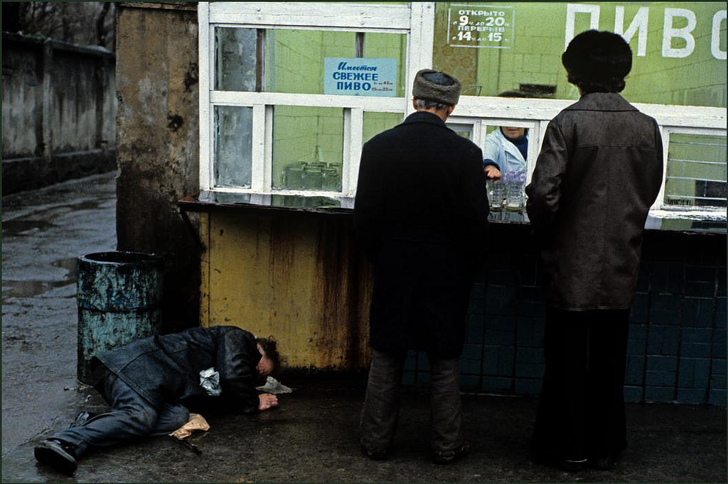 #7 Two men buying beer from a street kiosk in the rain whilst another lays drunk on the ground.
