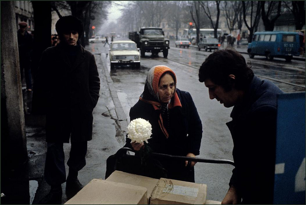 #32 A woman trying to sell a flower to a man unloading packages in the rain on a shopping street.