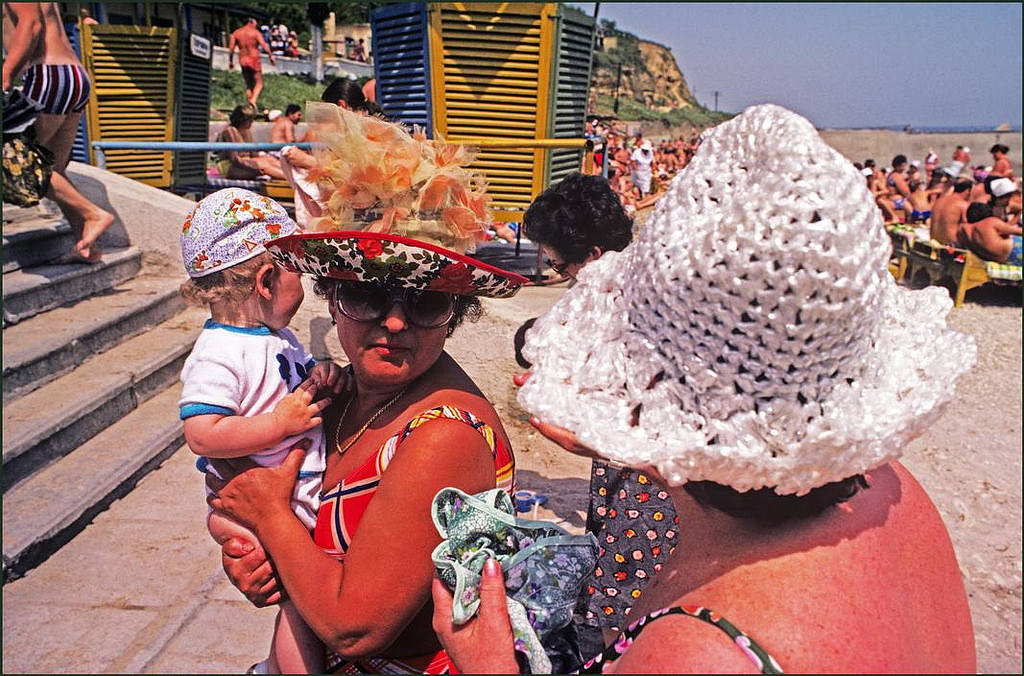 #14 A woman in an amazingly decorative sun hat holds her child whilst chatting to another lady on the beach.
