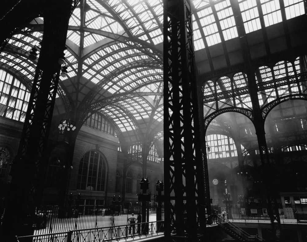 #18 Penn Station, Interior, Manhattan. Massive steel uprights in the center and to the left, with lighter steel tracery and windows above; men stand near stairway to trains.