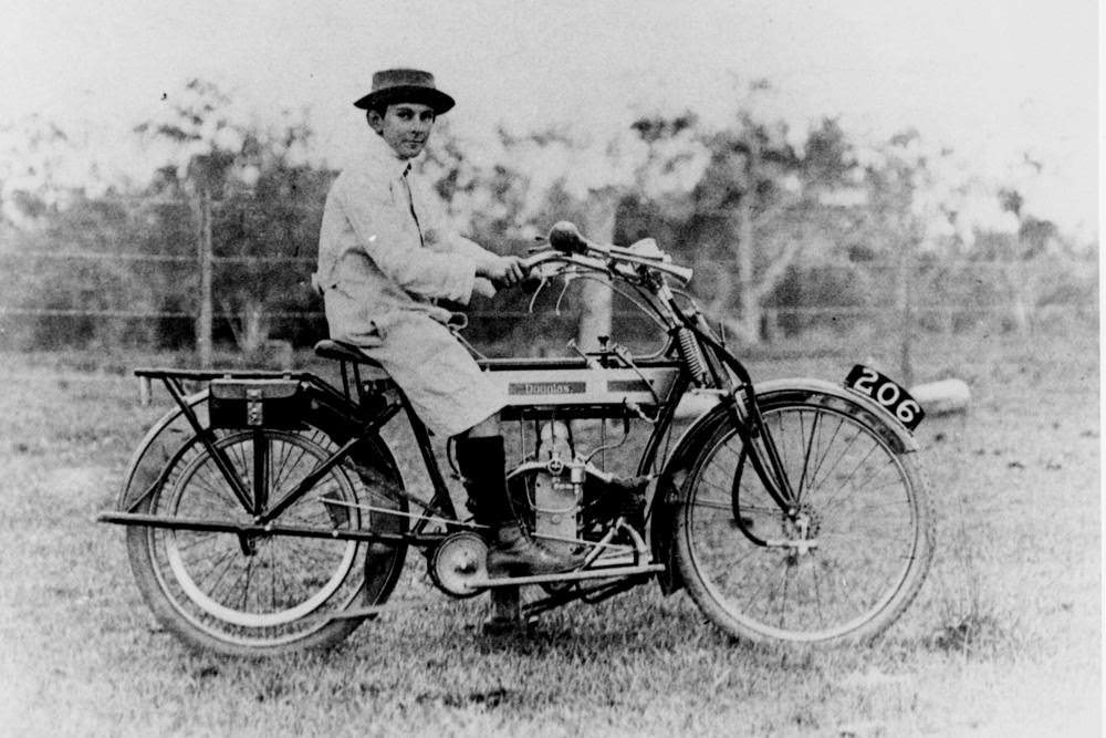 #68 A boy, wearing a long coat and a hat with the brim turned up, is sitting on a early model Douglas motorcycle, 1915.