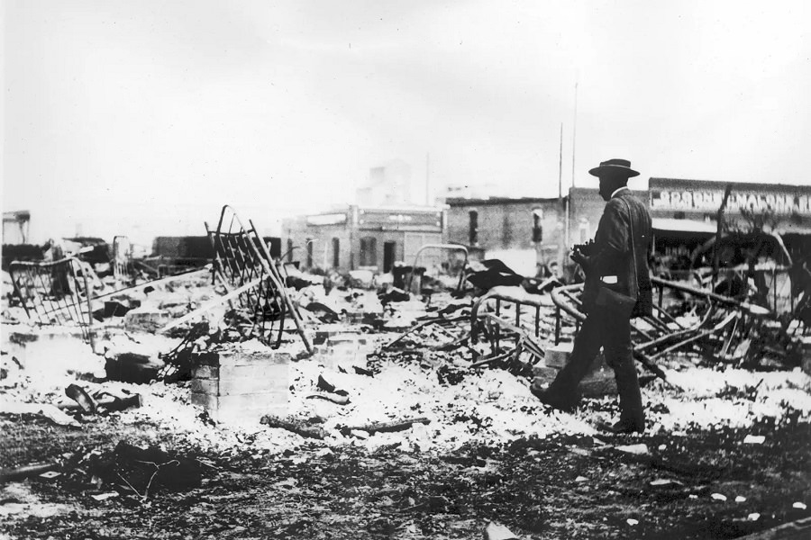 #19 A Black man with a camera inspects the skeletons of iron beds rising above the ashes of a burned-out block in Tulsa, June 1921.