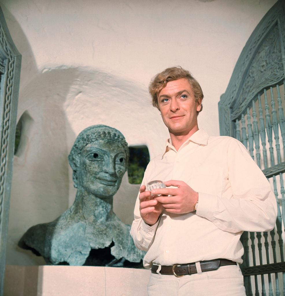 #1 Michael Caine stands next to a bronze Etruscan bust, 1960s.