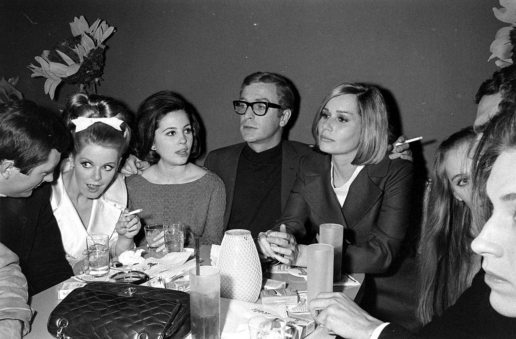 #64 Michael Caine smoking a cigarette and dining with friends, including the actress Sally Kellerman, 1966.