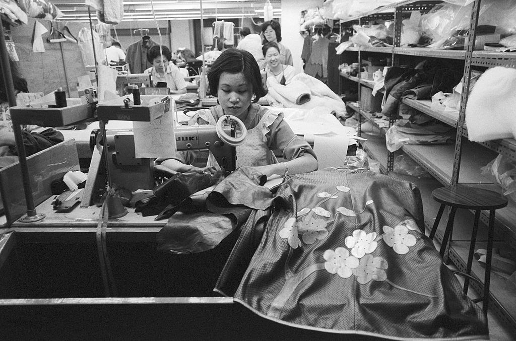 #31 A seamstress works in front of her sewing machine arranged perpendicular to a shelf. Hong Kong, 1981.