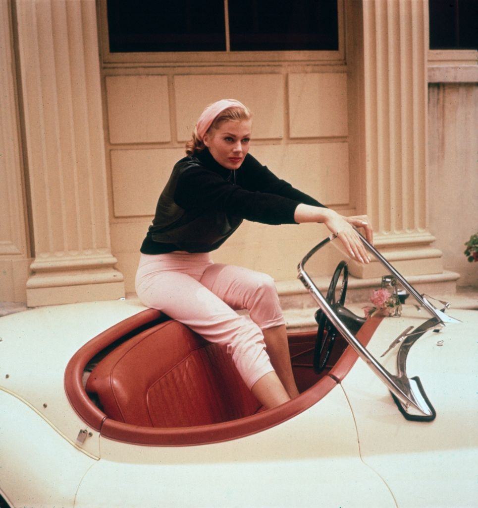 #33 Anita Ekberg sitting on the trunk of a cream convertible with her feet in its red leather interior, 1955.