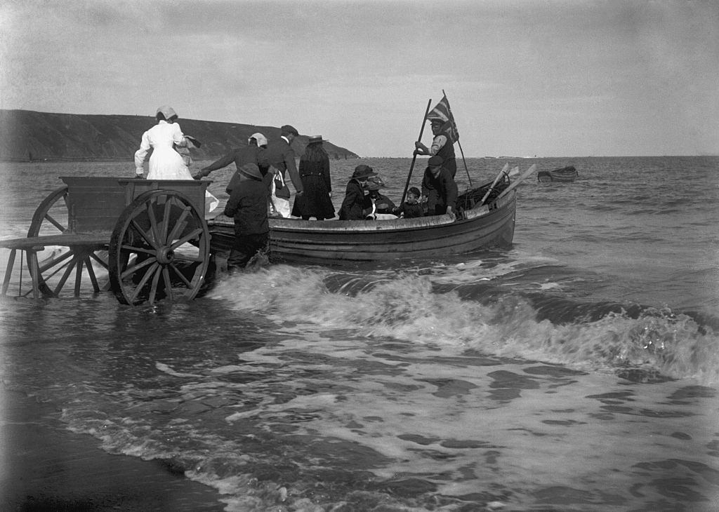 #13 Smartly dressed family group being helped from a two wheeled cart into a coastal rowing boat on an English beach circa 1900.