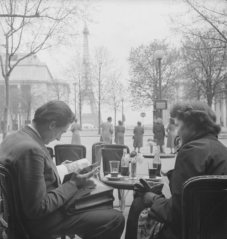 #13 An unidentified couple sit at a sidwalk cafe near the Eiffel Tower and read. Each has a bottle of Coca-Cola in front of them.