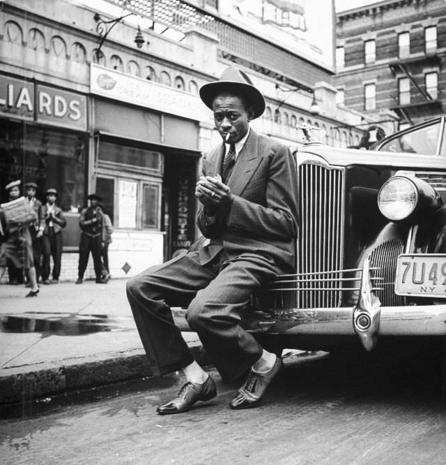 #10 Baseball player Satchel Paige, lighting his cigarette while sitting on the front bumper of a large car, 1941.
