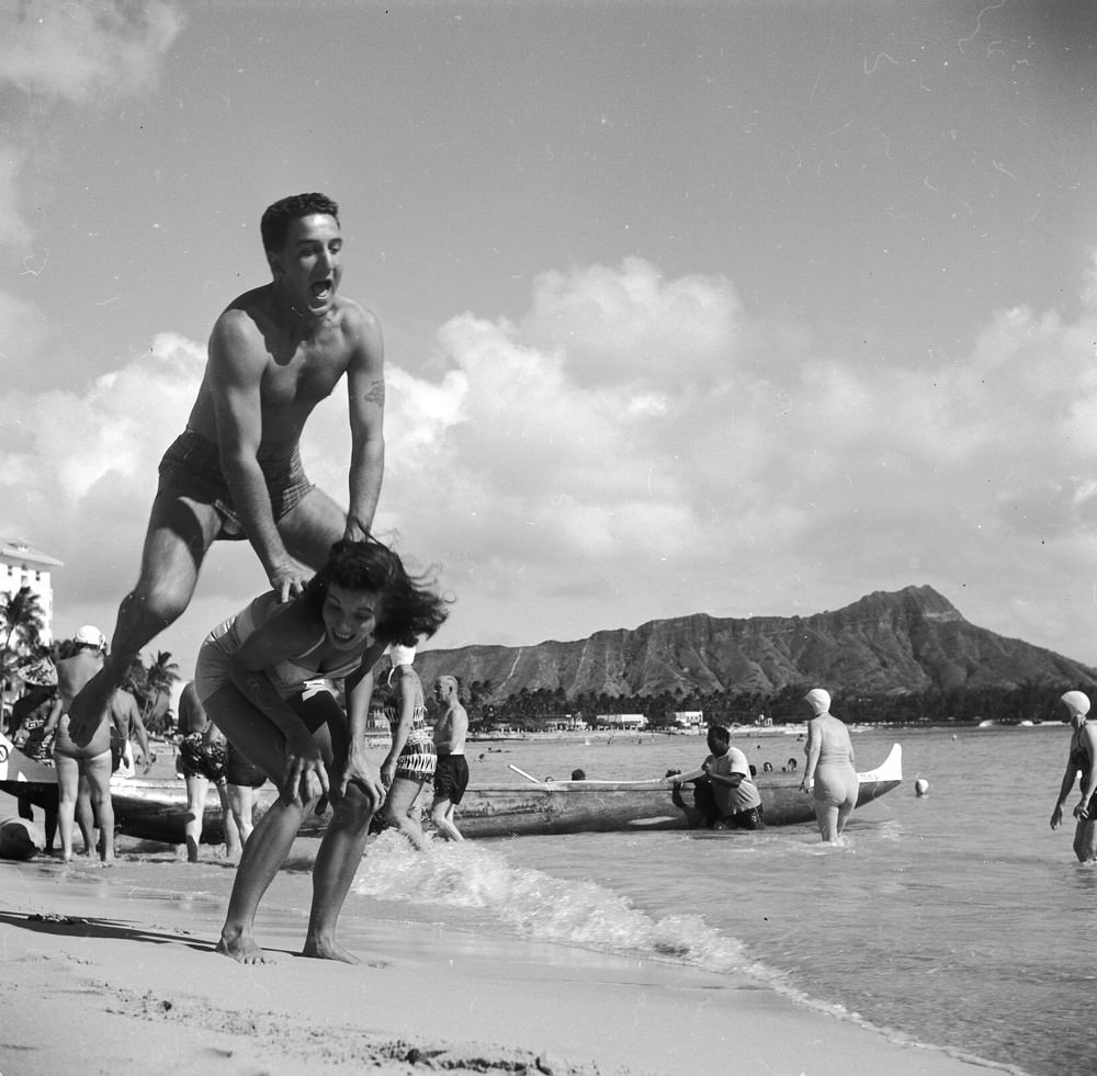 Bert and Ellie Lang, a young American couple on their honeymoon in Hawaii play leap-frog on Honolulu’s Waikiki beach. In the background is the extinct volcanic crater of Diamond Head, circa 1955.
