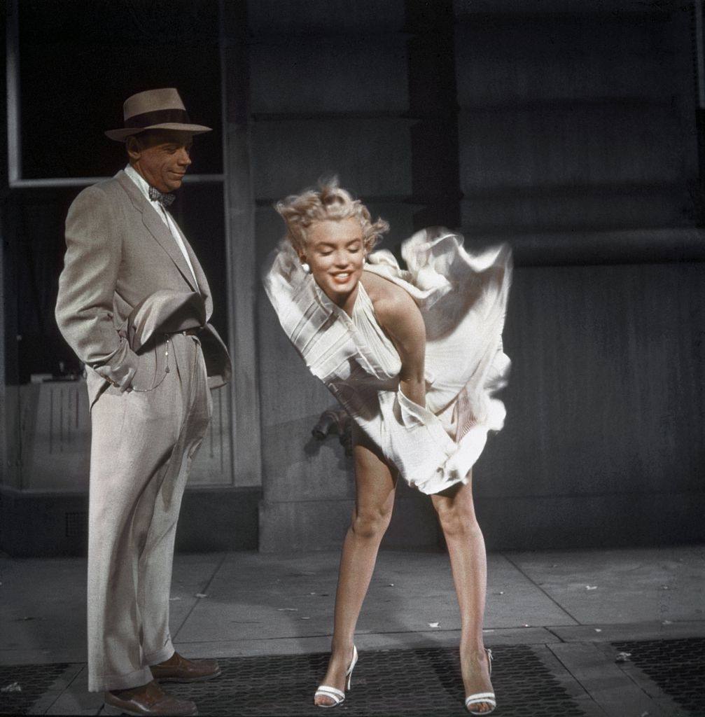 Marilyn Monroe standing over a subway grate with her white dress blowing and co-star Tom Ewell looking on during the filming of “The Seven Year Itch”, 1954.