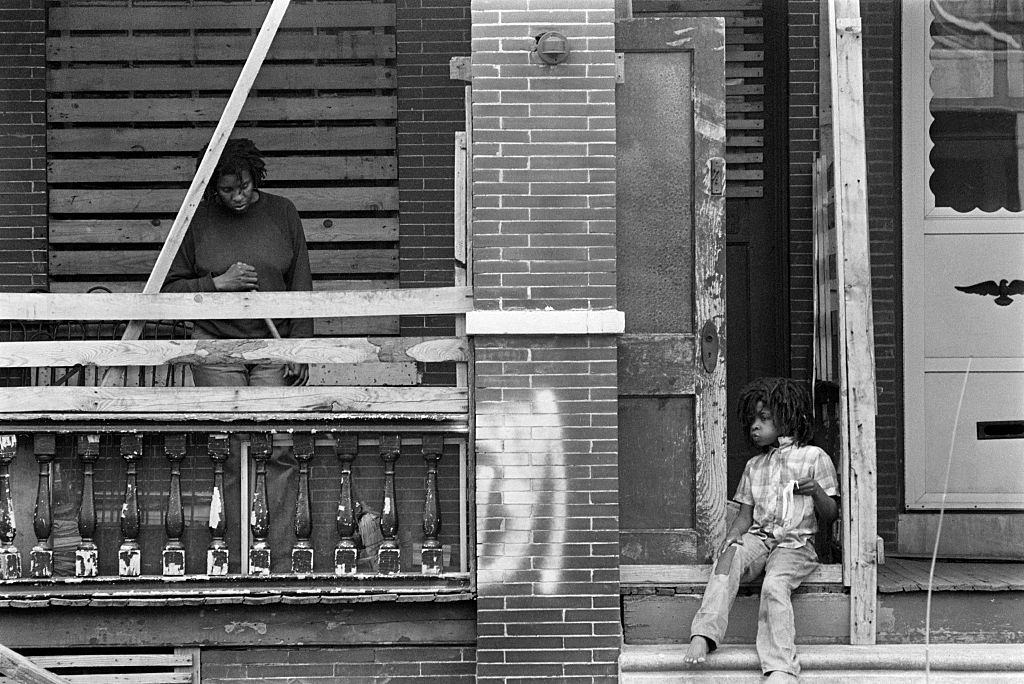 #2 A woman cleans her porch while a child in dreadlocks sits on the steps eating lunch.
