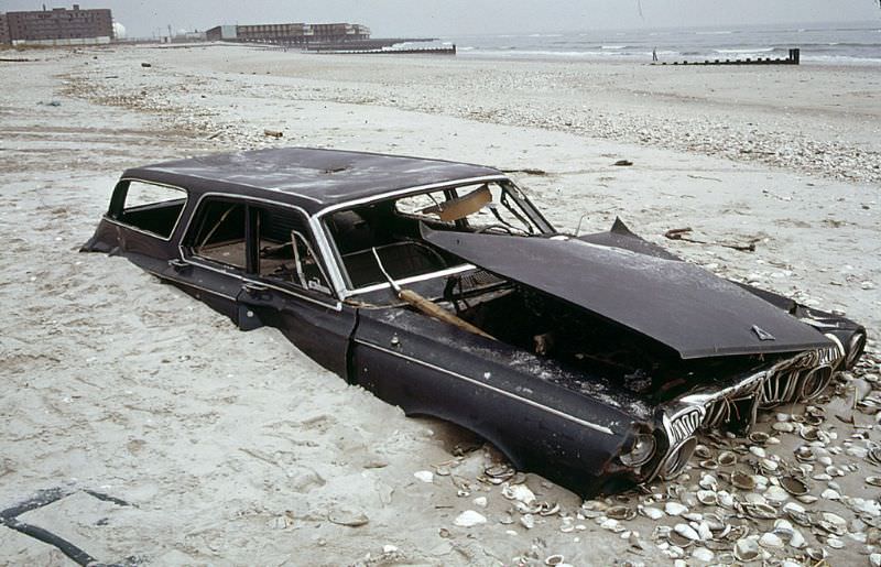 #21 Half buried early 1960s Dodge Polara station wagon on the beach at the ocean side of Breezy Point in Queens, an abandoned apartment building at left distance, 1973