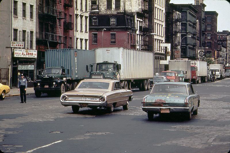 #19 A 1960s Ford Galaxie and a battered early 1960s Plymouth Valiant cruise along Canal Street with peeling pavement, 1973