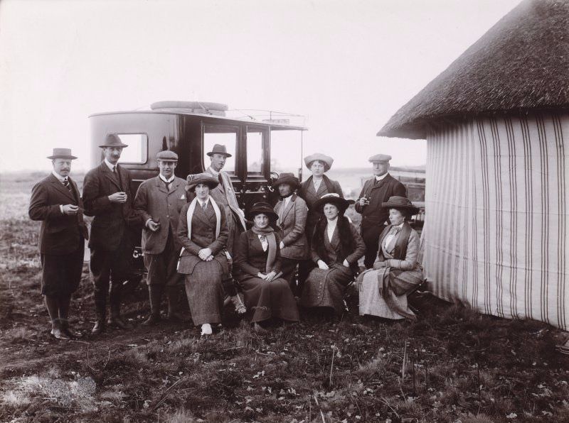 #65 Group photograph showing five men and five ladies beside a large car, outside what appears to be a thatched striped tent. Some of them are members of the Wallace family of Duchal House.
