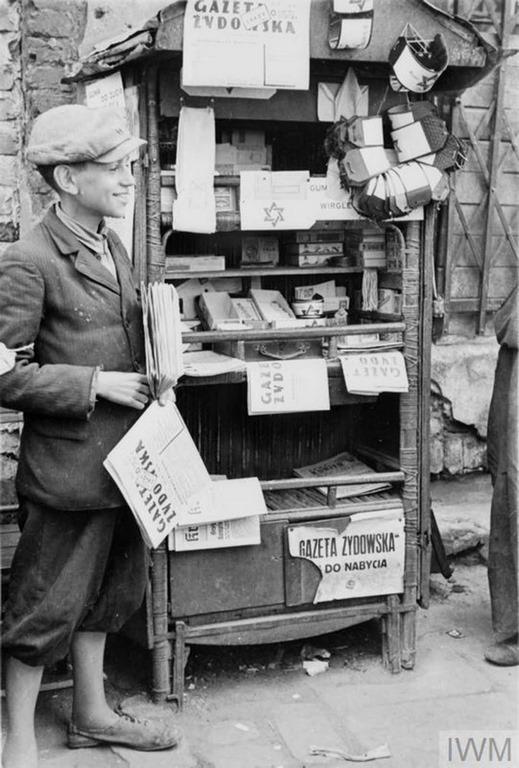 #12 A young and cheerful seller of newspapers and armbands running his stall in the street of the ghetto (possibly Muranowski Square). The title of the newspaper for sale is “Gazeta Żydowska – Jewish Gazette”. He also advertises Wrigley’s chewing gum for sale, mispelled as Wirgley’s.