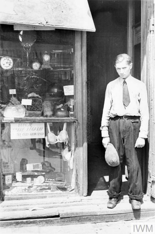 #13 A young man in the doorway of a shop in the ghetto. Note he has taken his hat off to comply with the German order to remove headwear in the presence of German personnel. The shop offers fresh eggs, sweets and watches. The sign in the window reads – “I buy old watches for top prices”.