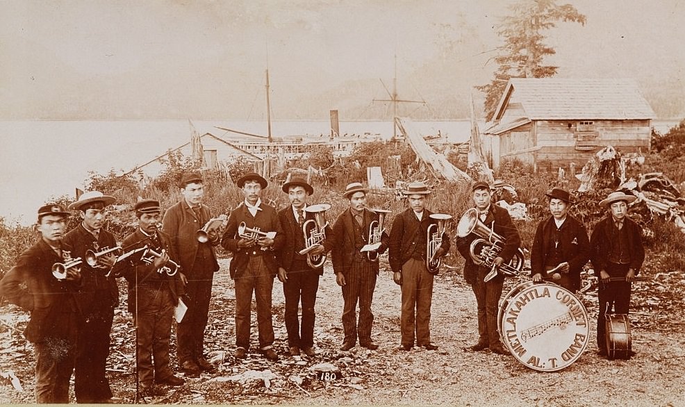 #8 Members of the Metlakahtla Cornet band, a group of Native American brass musicians, pose for a picture next to buildings by the shore in an Alaska town, ca. 1892.