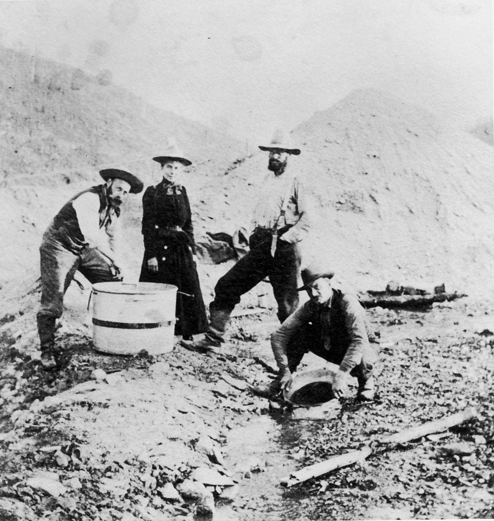#49 Three men and a woman standing and crouching around a riverbed while panning for gold during the Klondike gold rush, Alaska, 1895.