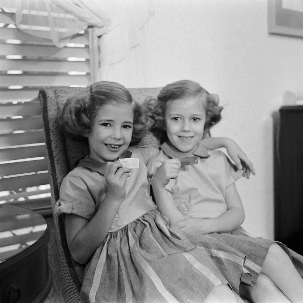 #10 Two sisters sitting in a chair in their home after an atomic war drill with their family, holding up the identification tags they wear around their necks, March 1954.