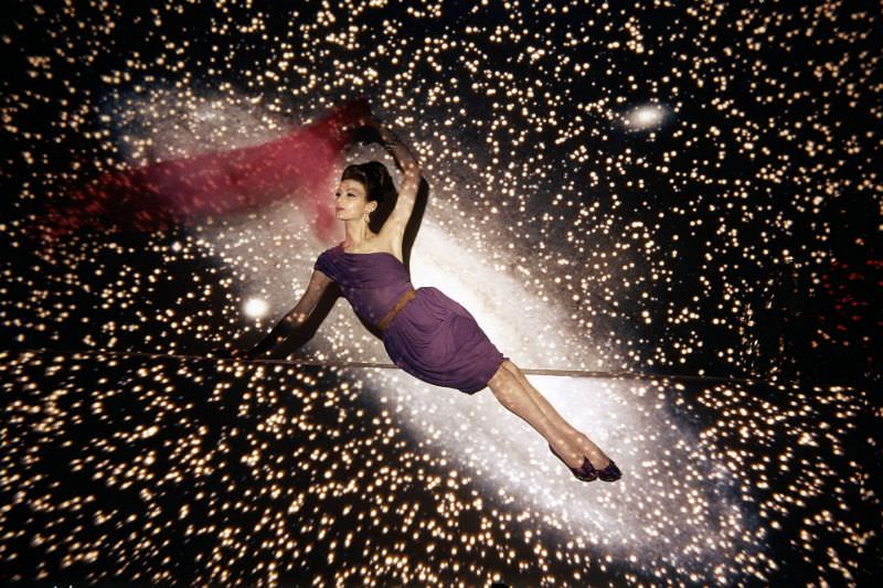 #31 Floating against a backdrop of the galaxy, Isabella is wearing violet short evening dress with pleats which drape across one shoulder by Maurice Rentner, 1960