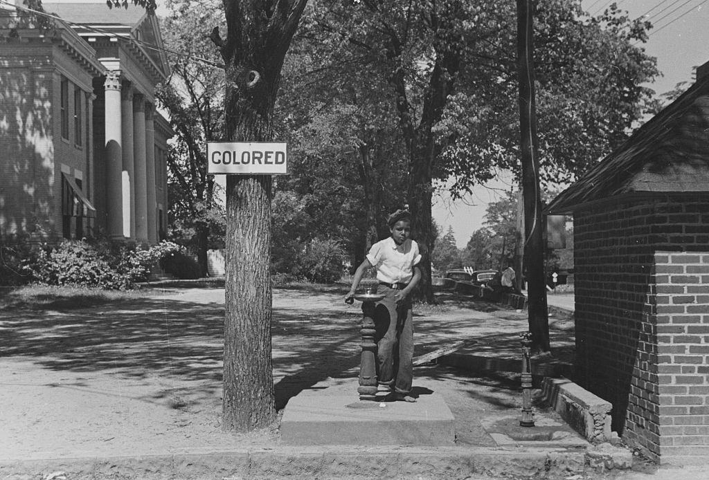 #4 African American child using a water fountain, the tree next to the fountain has a sign attached that reads ‘Colored’ in order to enforce Jim Crow laws