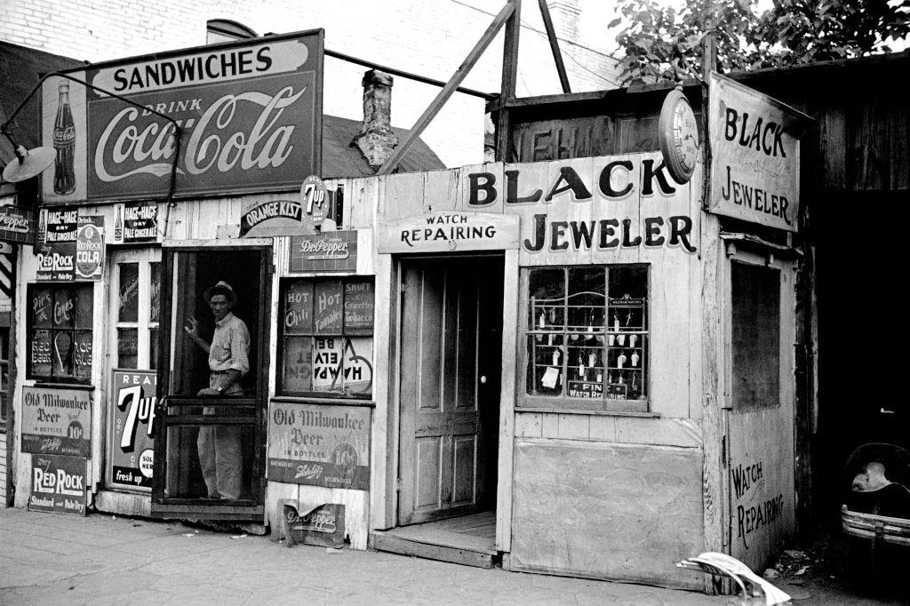 #5 Shack-like Black Jeweler shop next to a small food store covered with beverage ads in a slum section of the city of Atlanta, 1938.