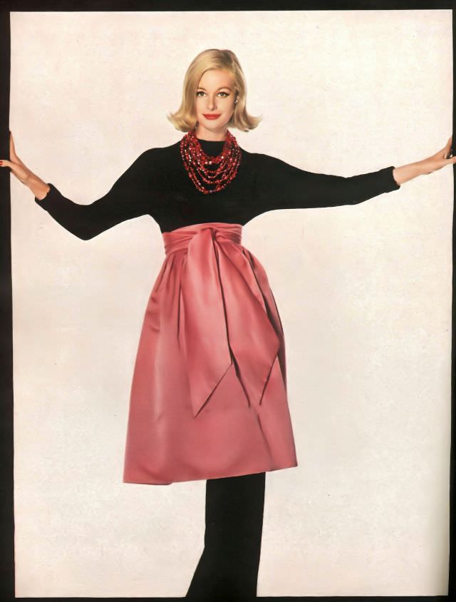 #37 Monique Chevalier is wearing a bright pink skirt-sized apron in satiny cotton; the streamers wrapped high on the waist of a toe-length black sweater, Vogue. December 1958