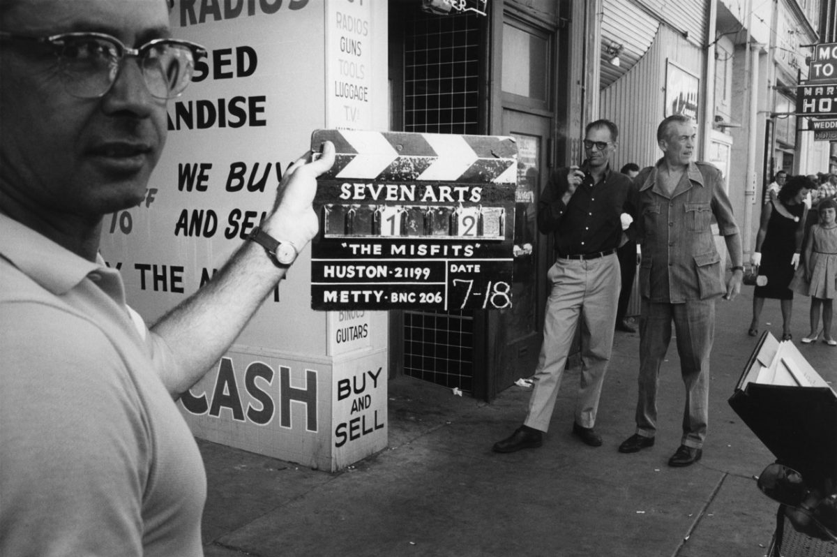 #20 Clapperboard is marked scene 1 take 2 on the first day of shooting. Director John Huston and author Arthur Miller watching in the background.