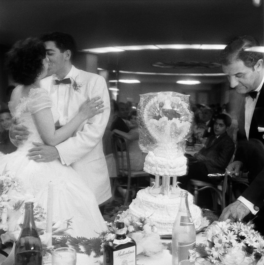#51 A newlywed couple kisses at their wedding reception, as their wedding cake is cut in Brooklyn Heights, March 1985.