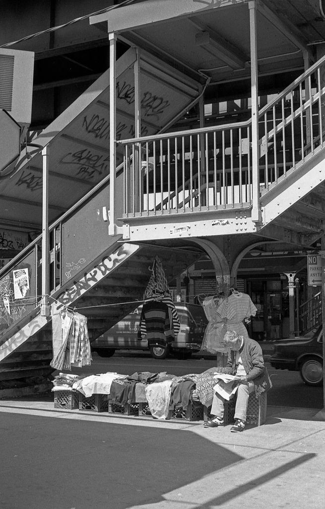 #14 A vendor selling clothes next to the staircase leading to the 103rd Street elevated subway station in the Corona neighborhood. Queens, New York, 1990.
