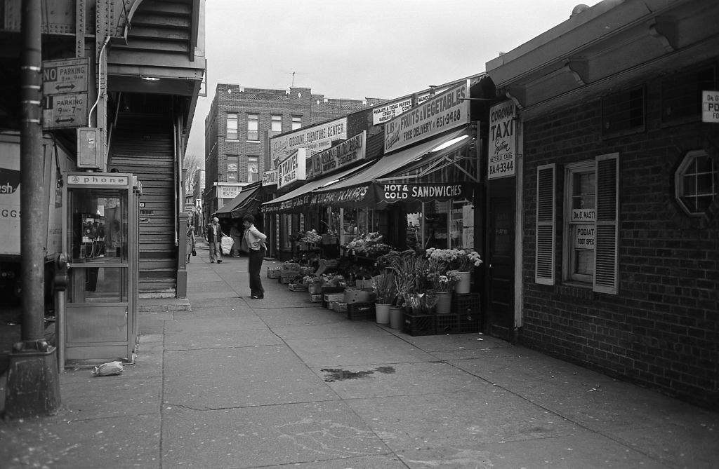 #8 A lady exits a fruit and vegetable stand below the elevated subway line on Roosevelt Avenue in the heart of Corona, Queens. 5th November 1974