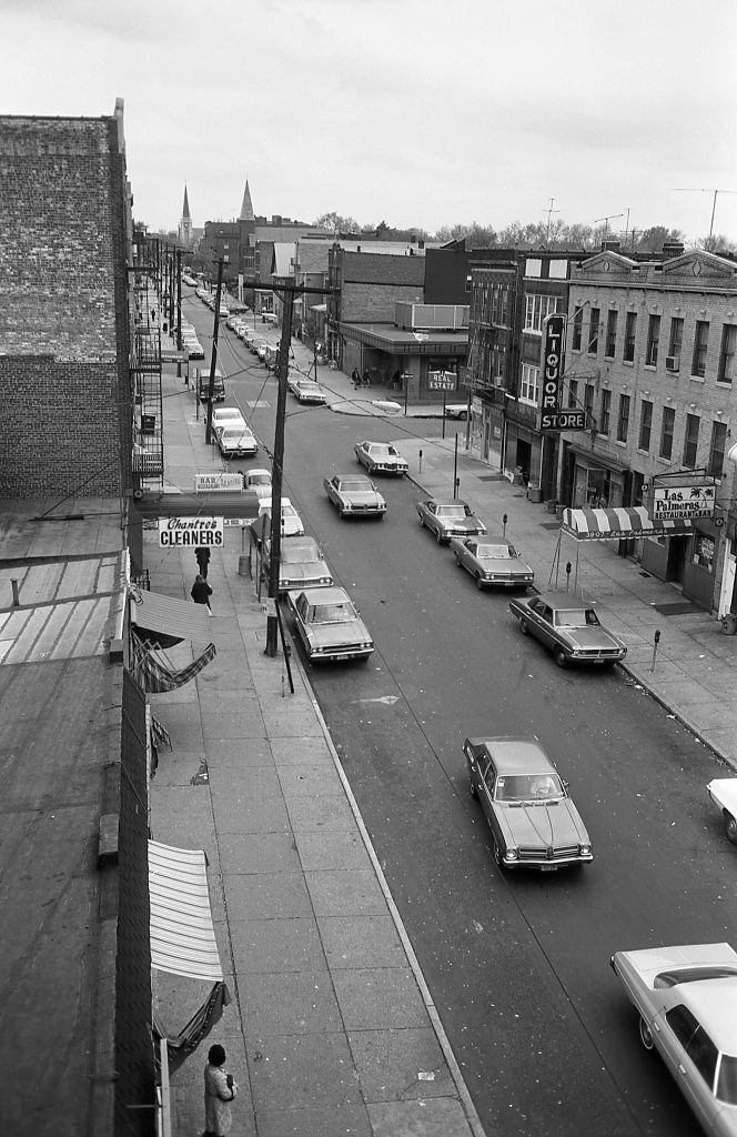 #18 Elevated subway line of a street lined with small businesses in Corona, Queens, 1974.