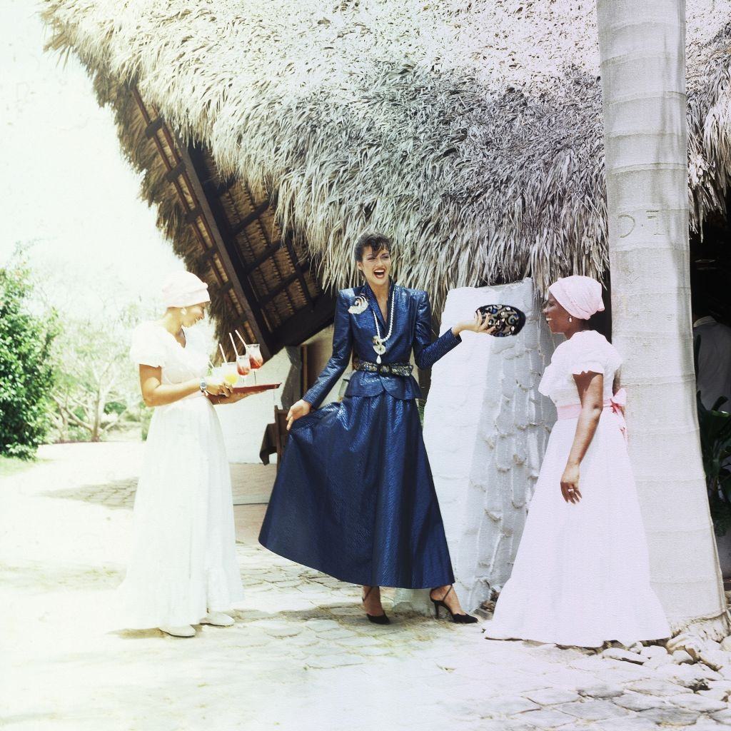 #25 Janice Dickinson in The Dominican Republic wearing a long blue skirt and matching jacket, 1979.