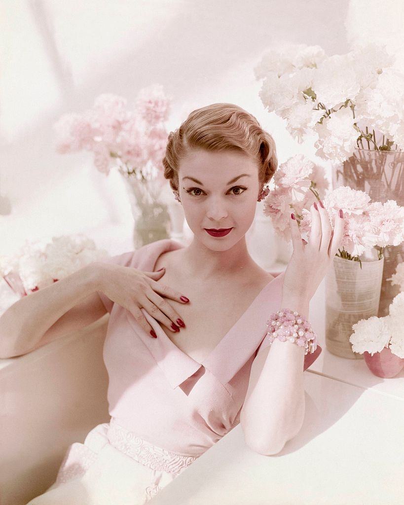 #17 Jean Patchett seated among vases of pink and white carnations wearing pink open neck linen dress with wide lapel collar and matching white with pink brocade skirt. Vogue 1950