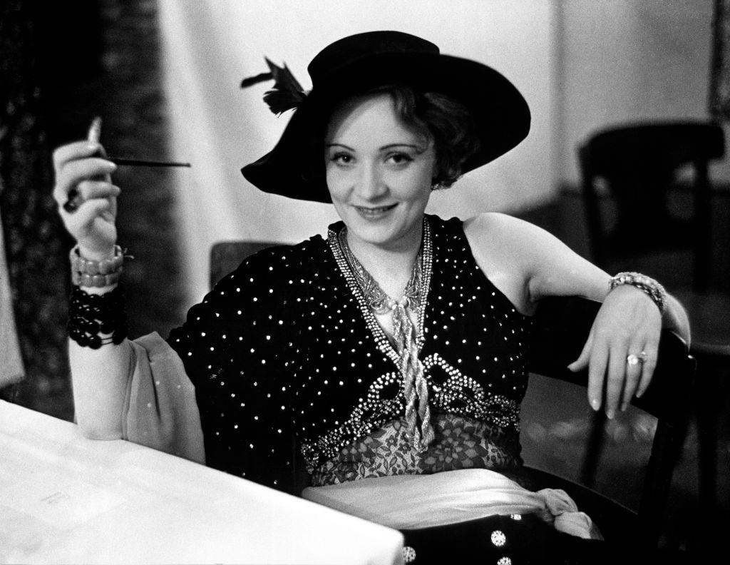 #52 Marlene Dietrich in evening dress and hat, smiling while sitting at table alone during Pierre Ball.