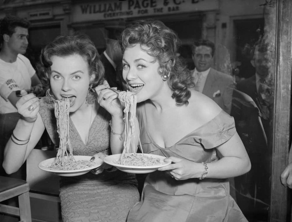 #8 Mirv Arvinen (Miss Finland 1955) and Soho’s Fair Queen Andria Loran (right) try their hand at scoffing a plate of pasta at the Soho Fair spaghetti-eating contest on Frith Street, Soho, London in 1956.
