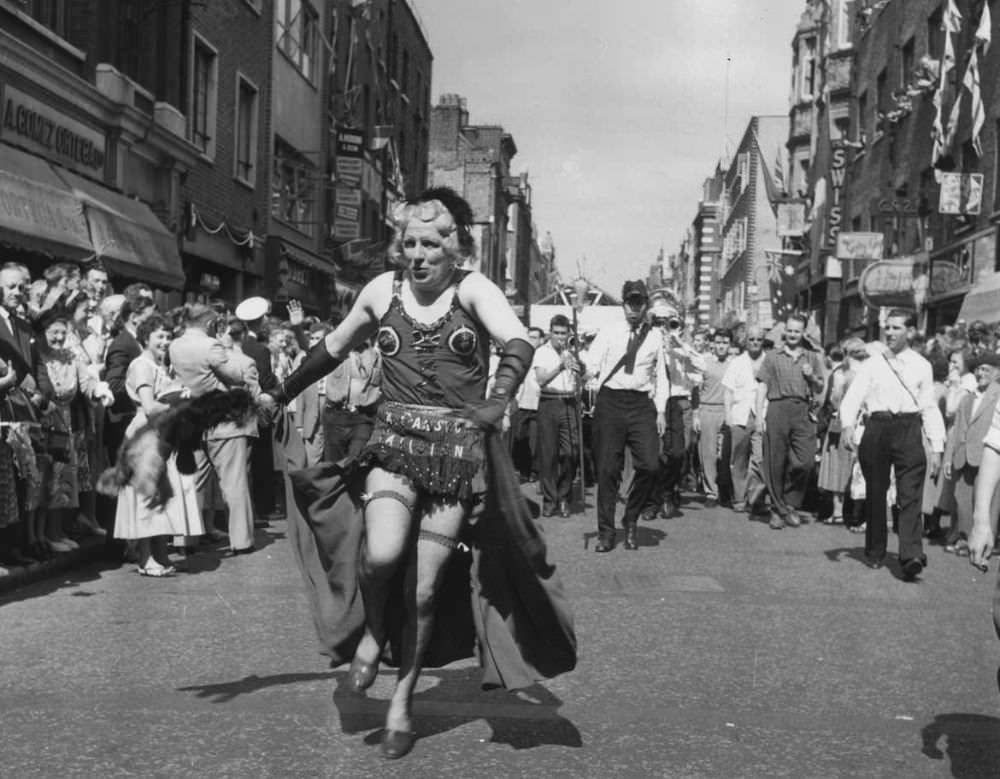 #11 A man in drag heads a carnival procession down Old Compton Street during the Soho Fair in 1956.