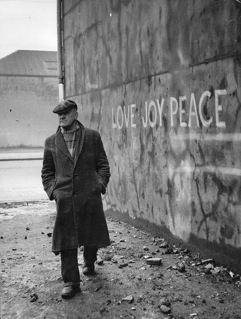 #46 Retired lion-tamer Alec Robinson walks past a Belfast wall scrawled with the slogan ‘Love – Joy – Peace’, 1955.